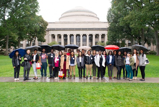 23 students stand under umbrellas in front of the MIT dome.