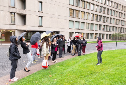 A group of students stand outdoors under umbrellas and listen to a guide while on a campus tour.