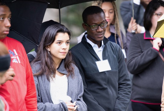A young man and a young woman listen to a guide as they enjoy a campus tour.