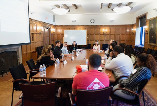 A group of students sit around a conference table with a panel of professors and professionals.