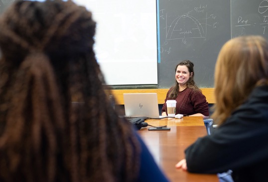 A seated female graduate student smiles as she addresses a conference table of program participants.