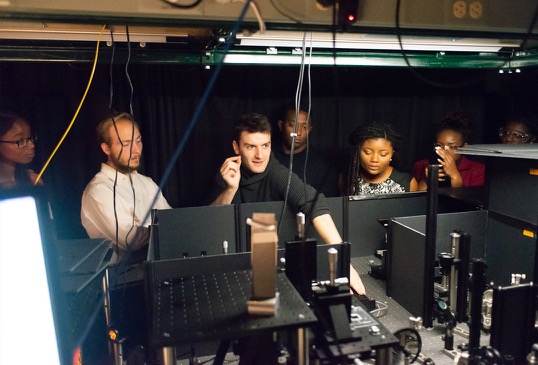 A male graduate student explains a laser set up to a group of program participants.