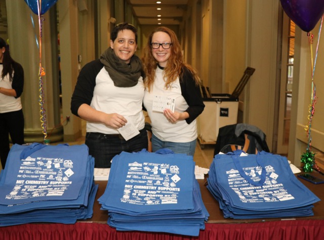 Two female staff members smile behind a table laden with custom tote bags.