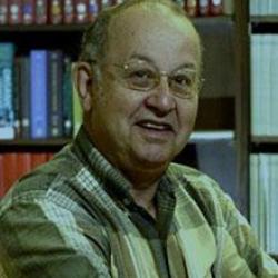 An elderly man smiles in front of a full bookshelf.