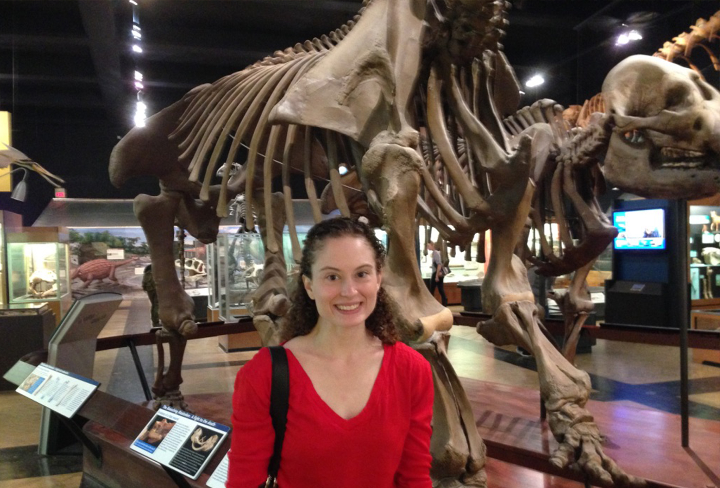 A young woman stands in front of a giant dinosaur skeleton.