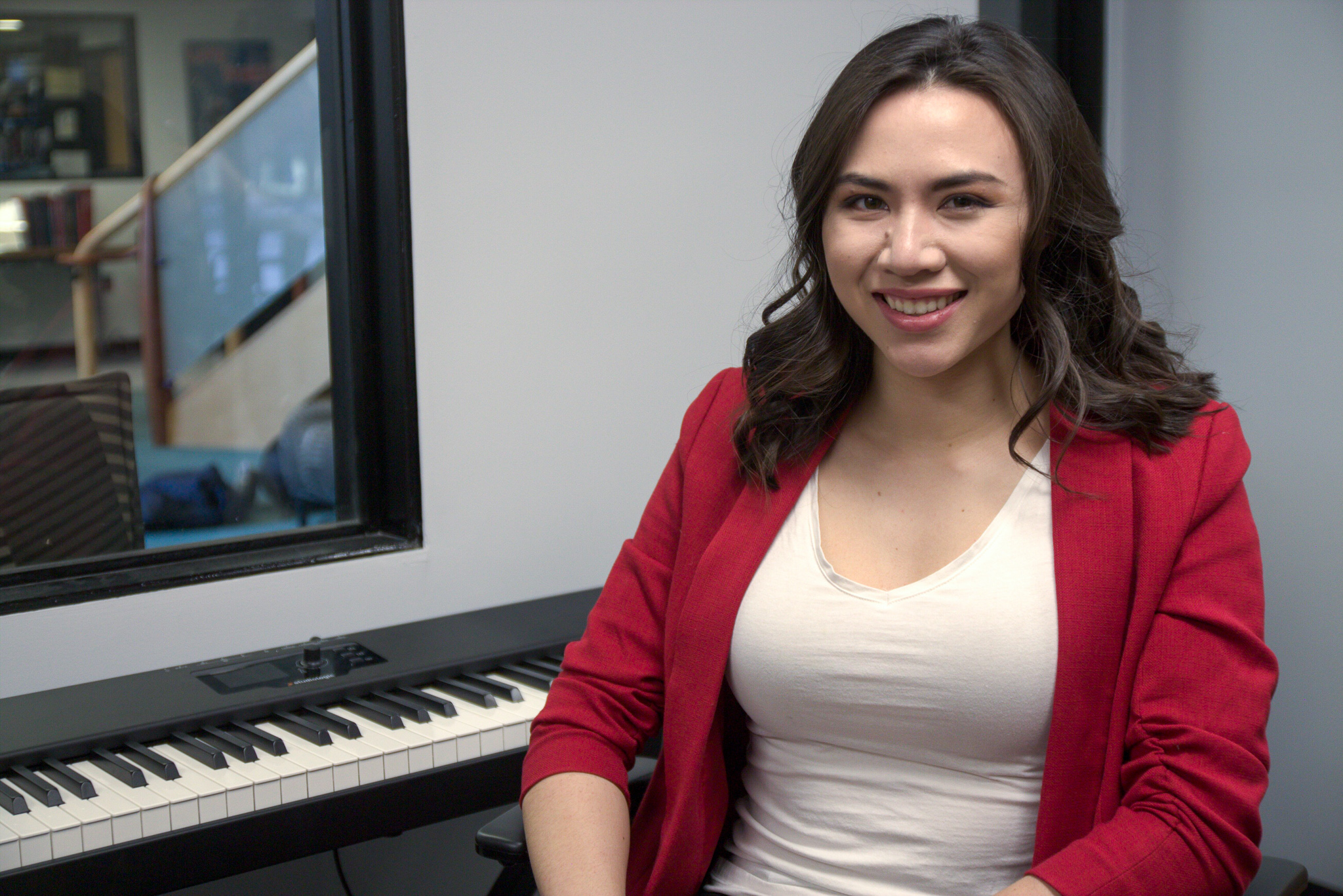 A young woman with brown hair sits smiling in front of a piano keyboard.