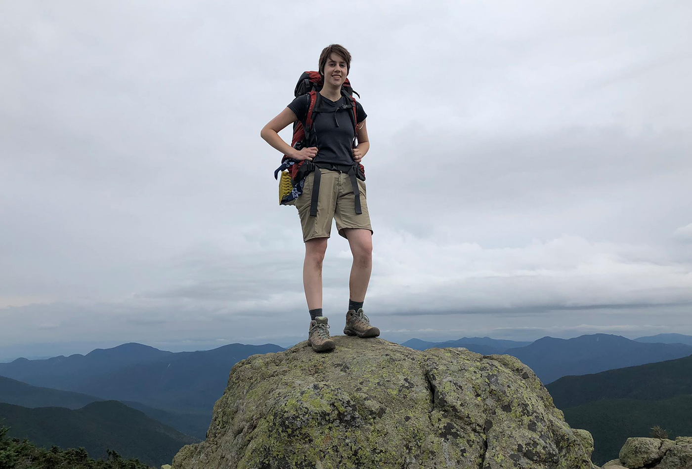 A female graduate student in hiking gear stands on top of a mountain.