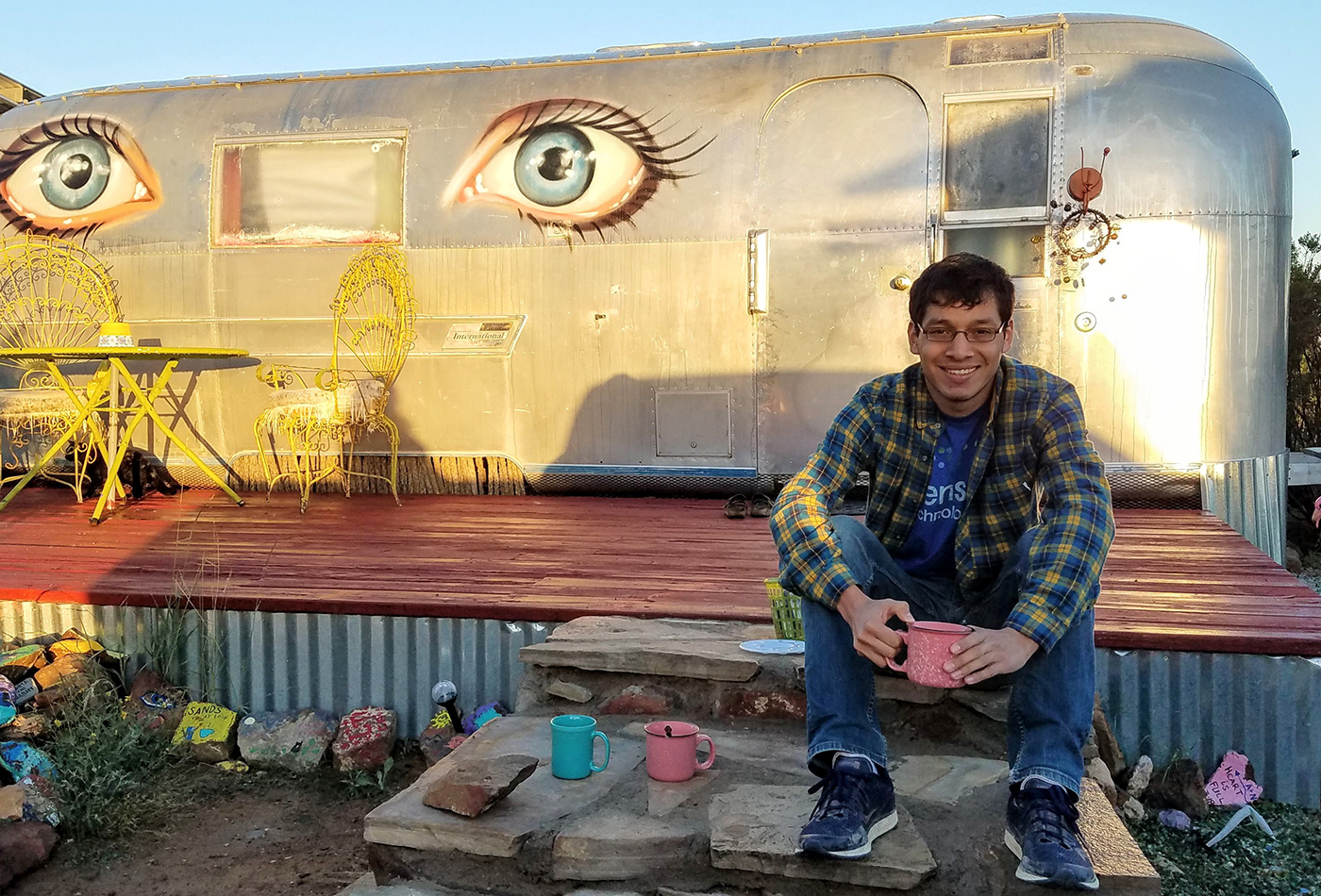 A male graduate student sits in front of an airstream trailer painted with two blue eyes.