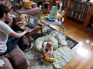 A man sits and works at a table with a baby at his feet.