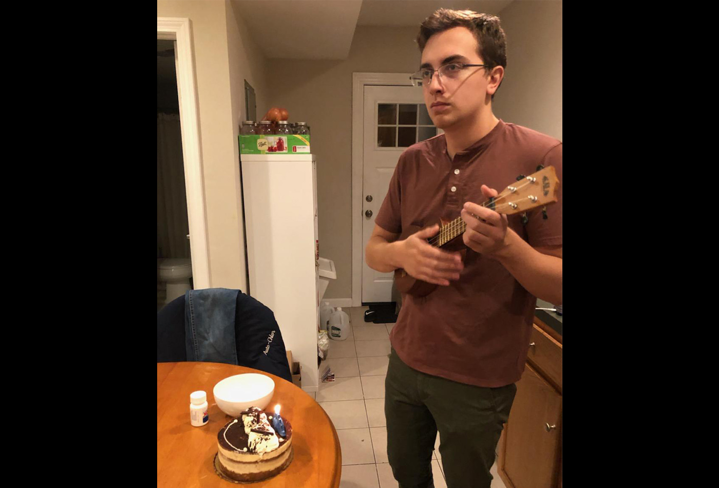 A male graduate student plays a ukelele in front of a cake with a lit candle on it.