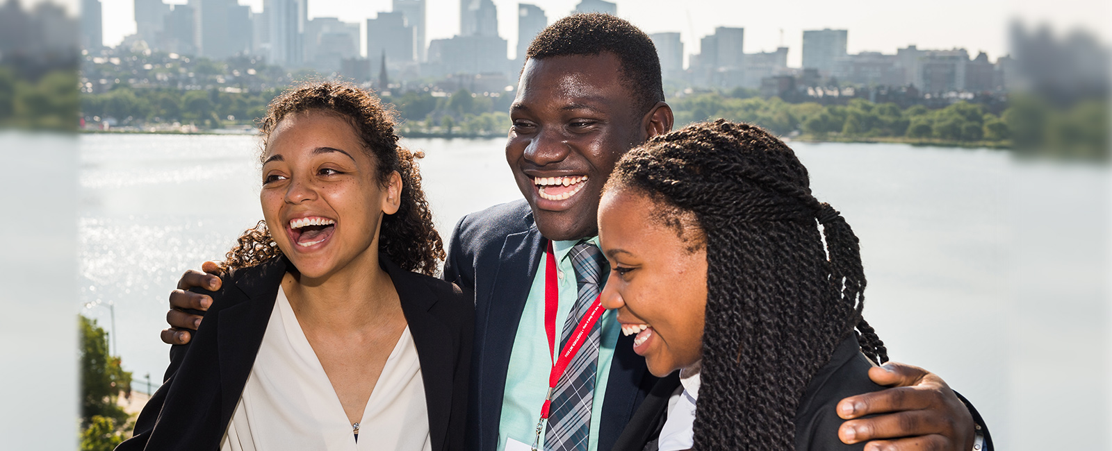 A trio of students of color smile in front of the Boston skyline.