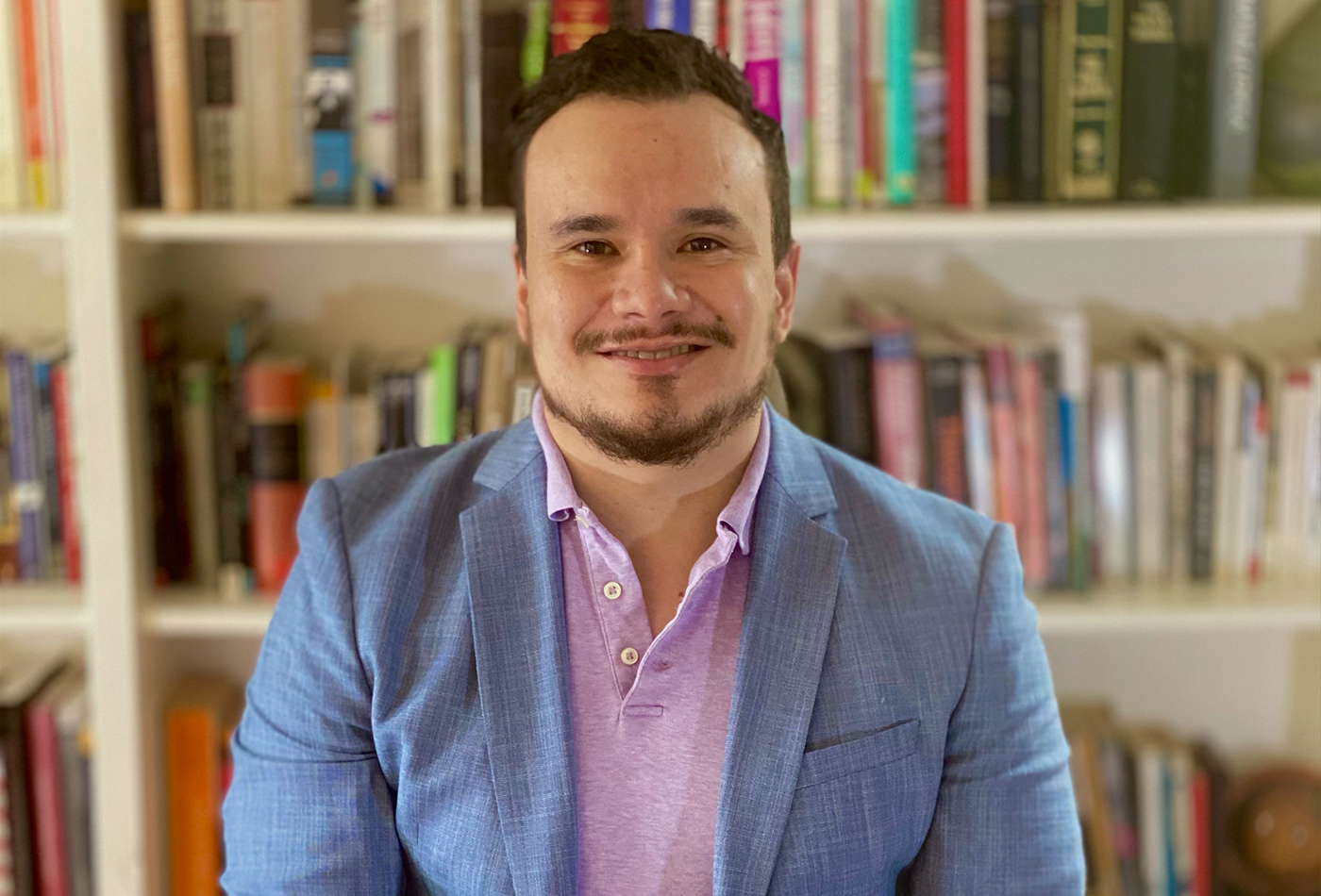A man in a blazer smiles in front of a bookshelf.