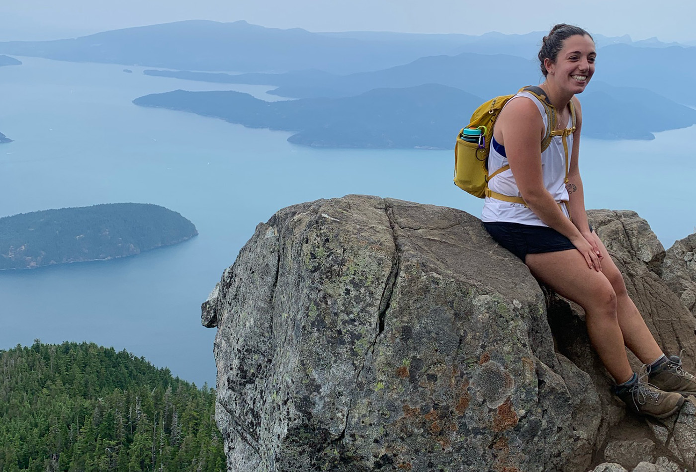 A graduate student smiles on a rock atop a mountain overlooking a lake.