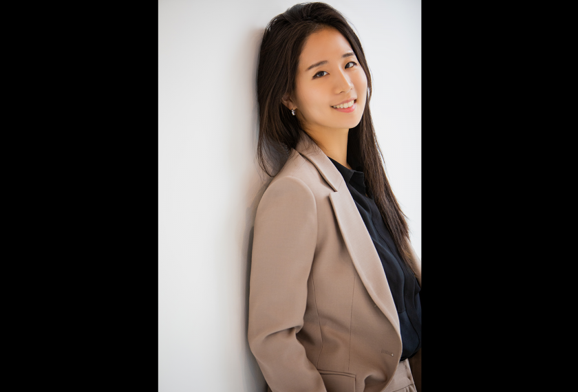 A person with long hair wearing a tan blazer leans against a wall and smiles