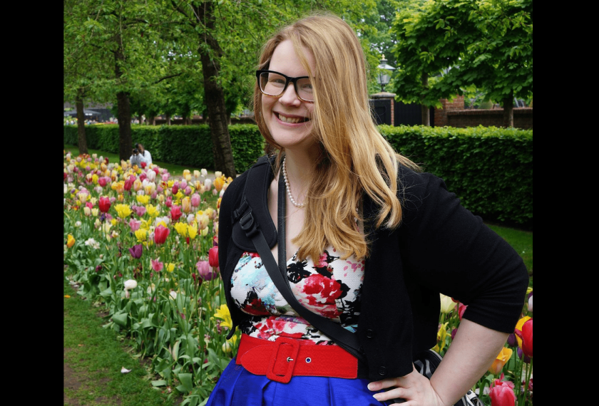 A person with long blonde hair and glasses smiles at the camera in front of a field of flowers