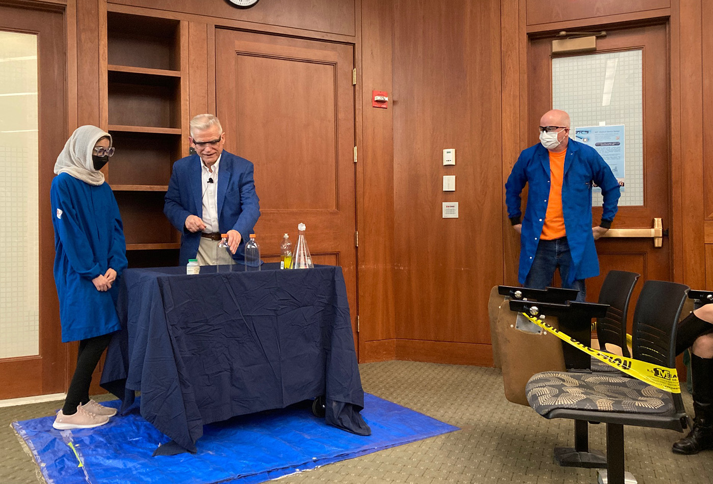 A young woman in a hijab, a white haired man, and a bald man in an orange shirt stand in a room, wearing lab coats.
