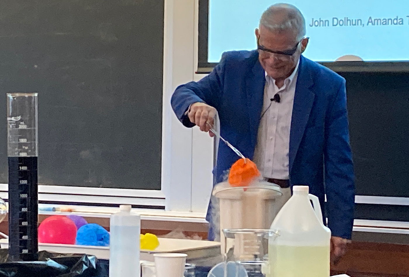 A white haired man in a lab coat pulls a shriveled balloon from a container full of dry ice.