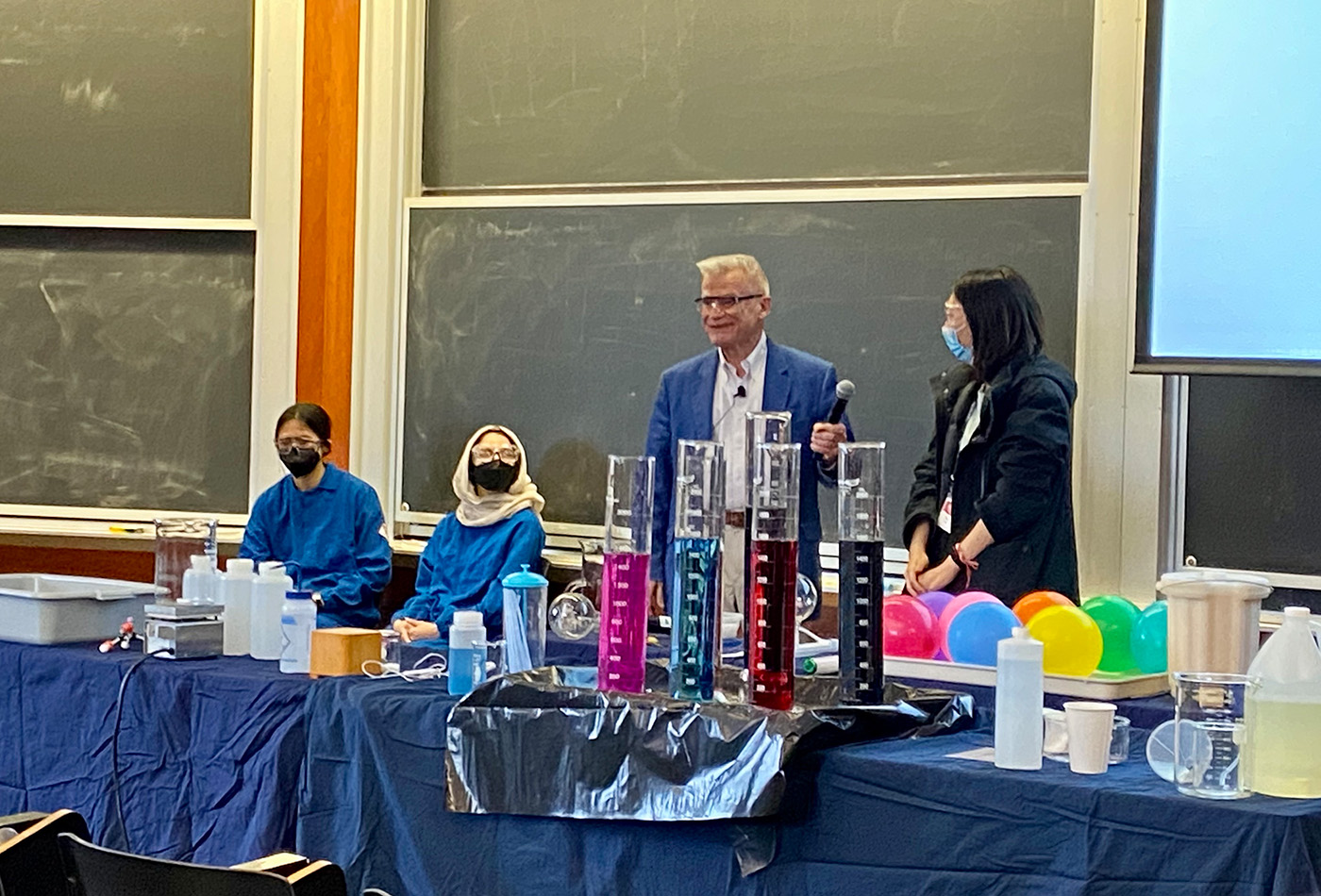 A white haired man stands beside a young woman with dark hair, in front of a table laden with various materials for science experiments.