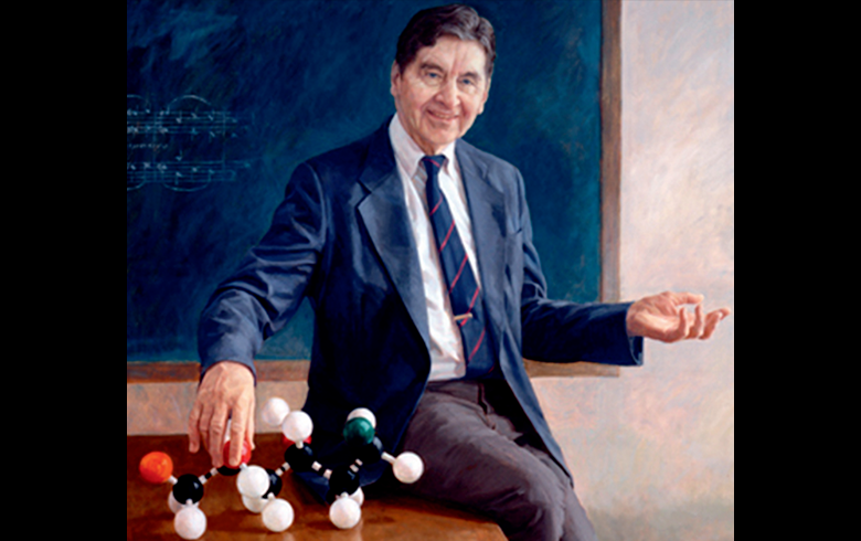 An old photo depicting a man in a blazer and tie sitting on a desk with a model of molecules.