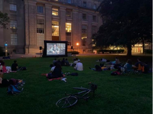Students sit on a sprawling lawn adjacent to a four story building watching a movie on an outdoor screen on a summer evening.