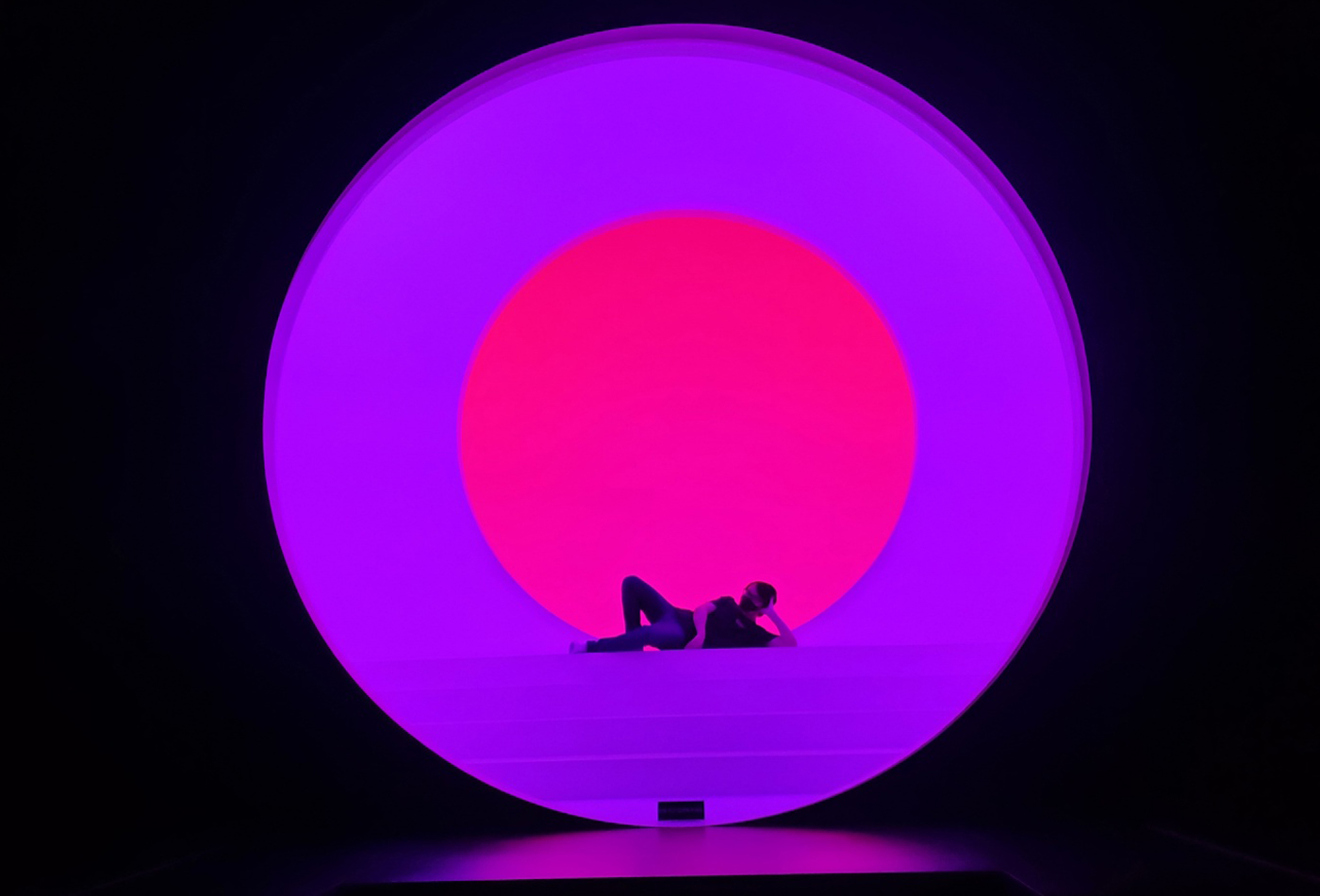 Michael Lee lays on his side, backlit by an art installation that is a lit purple circle with a smaller pink circle inside of it. Michael is a dark shadow in front of these two circles.