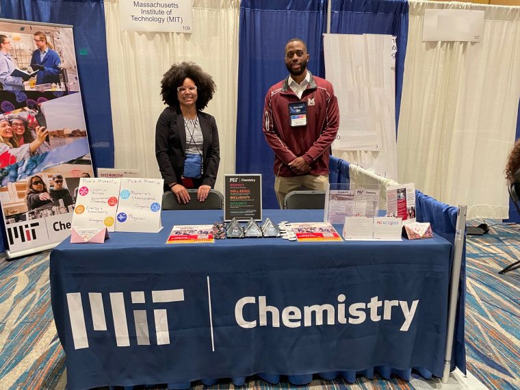 Kayla Storme and Andy Dorfeuille stand behind a booth covered with a blue tablecloth boasting the MIT Chemistry logo in a conference hall.