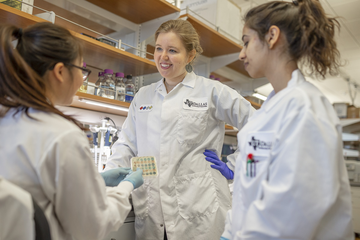 Nicole De Nisco stands in a lab between two female students, facing her. All are in lab coats.