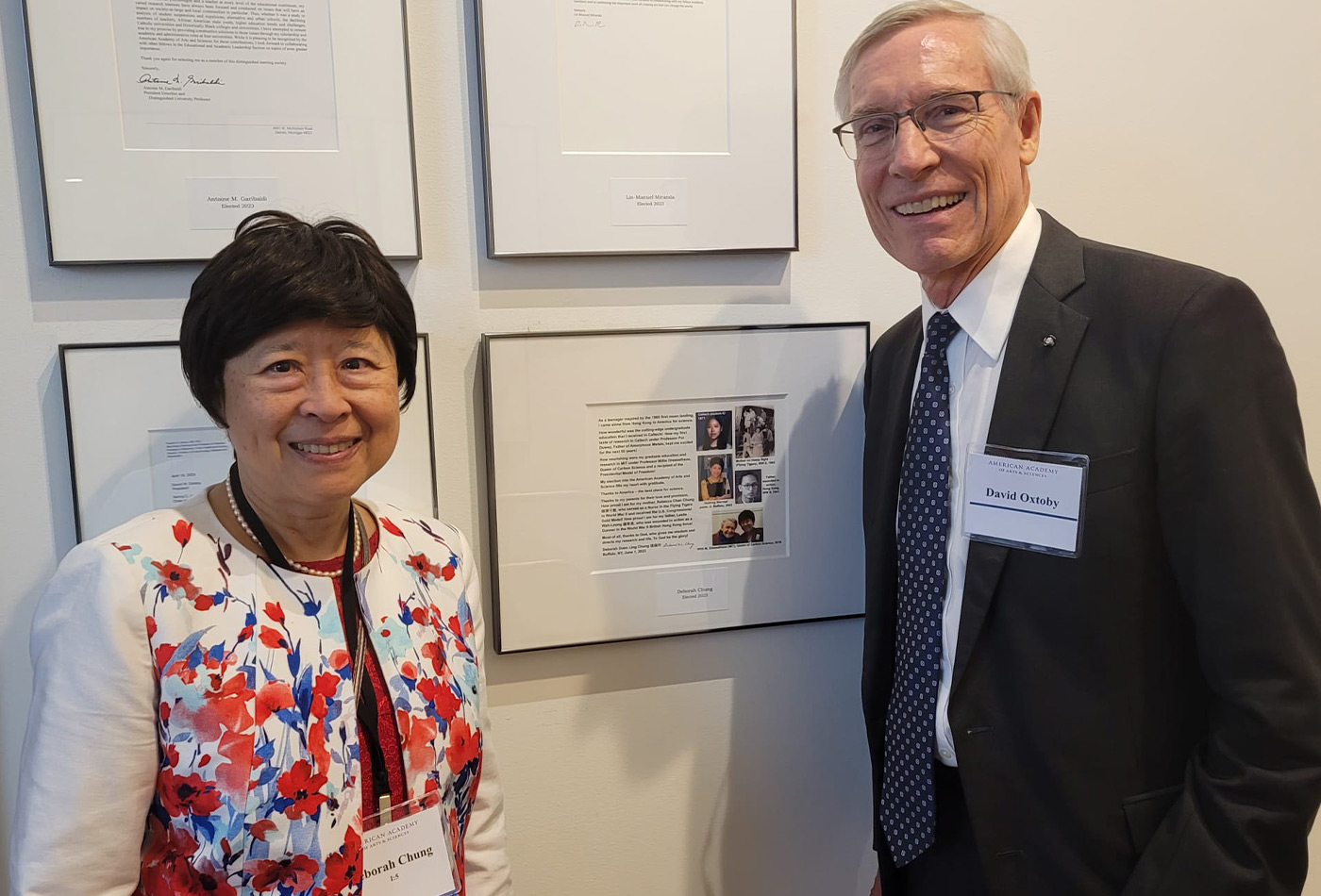 Deborah Chung smiles with David Oxtoby in front of a framed print out of Deborah's American Academy of Arts and Sciences induction letter that is hanging on the wall.