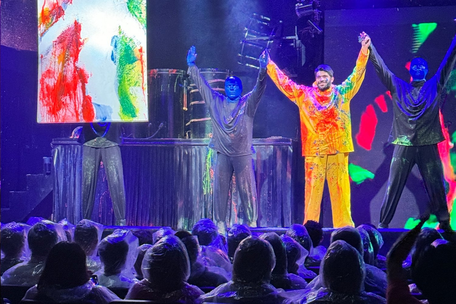 A student clad in a yellow rain suit splattered with paint stands between two men with blue heads and takes a bow on stage.