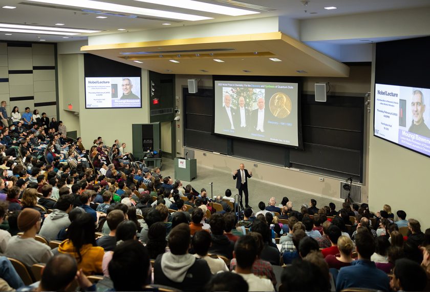 A man in a suit (Moungi Bawendi) speaks to a packed lecture hall, standing before three display screens.