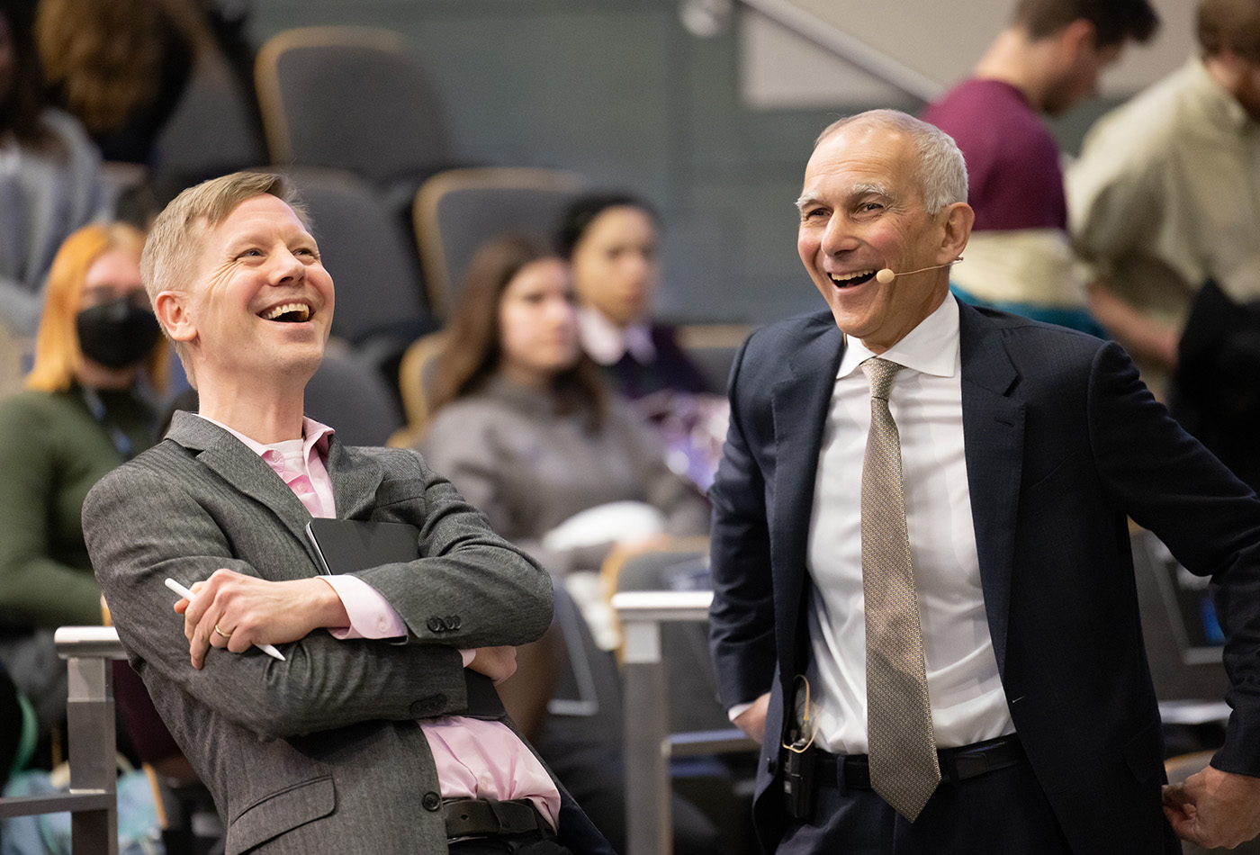 Two men wearing suits laugh together in a lecture hall.