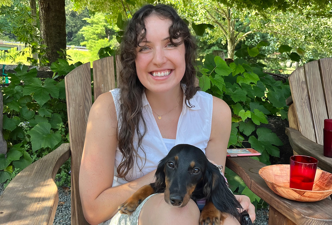 Katie Pelham sits on a wooden chair with a black longhaired daschund in her lap.