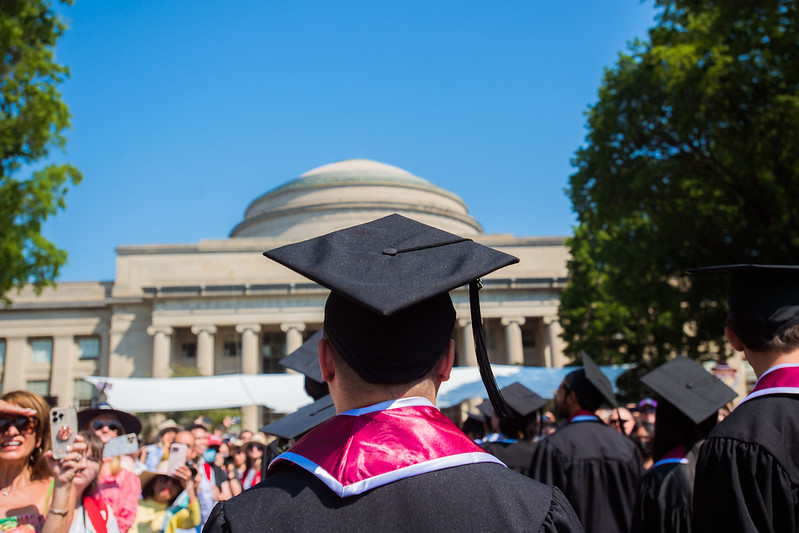 A student faces away from the camera wearing graduation regalia. They stand on Killian Court, looking toward the Dome.