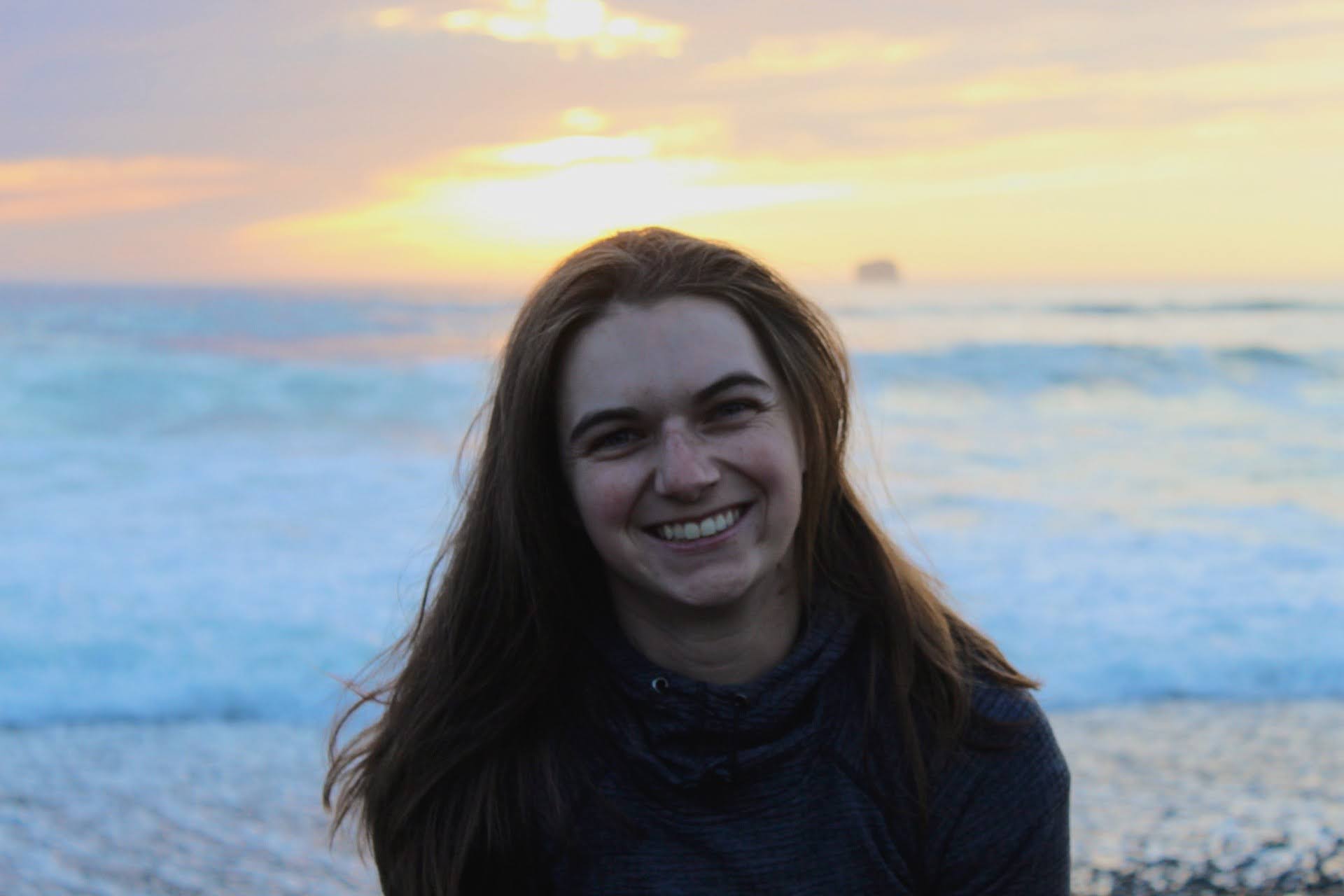 Sarah Quinn smiles in front of a sunset on a beach.