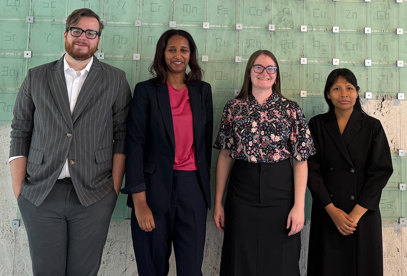 The four participants of the 2024 Future Faculty Symposium smile in front of a green glass sculptural installation of the periodic table of elements.