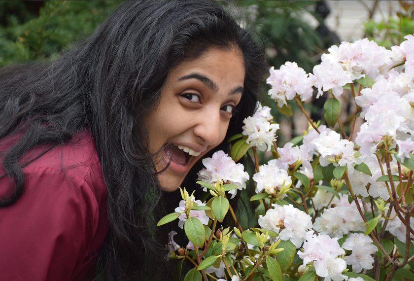 Maya Chattoraj smiles next to a bush blooming with pale pink flowers.