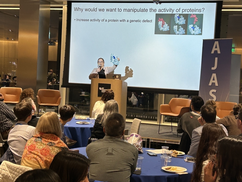 Photo of Professor Catherine Drennan delivering a keynote address from a podium while holding a model of molecules in her hand.