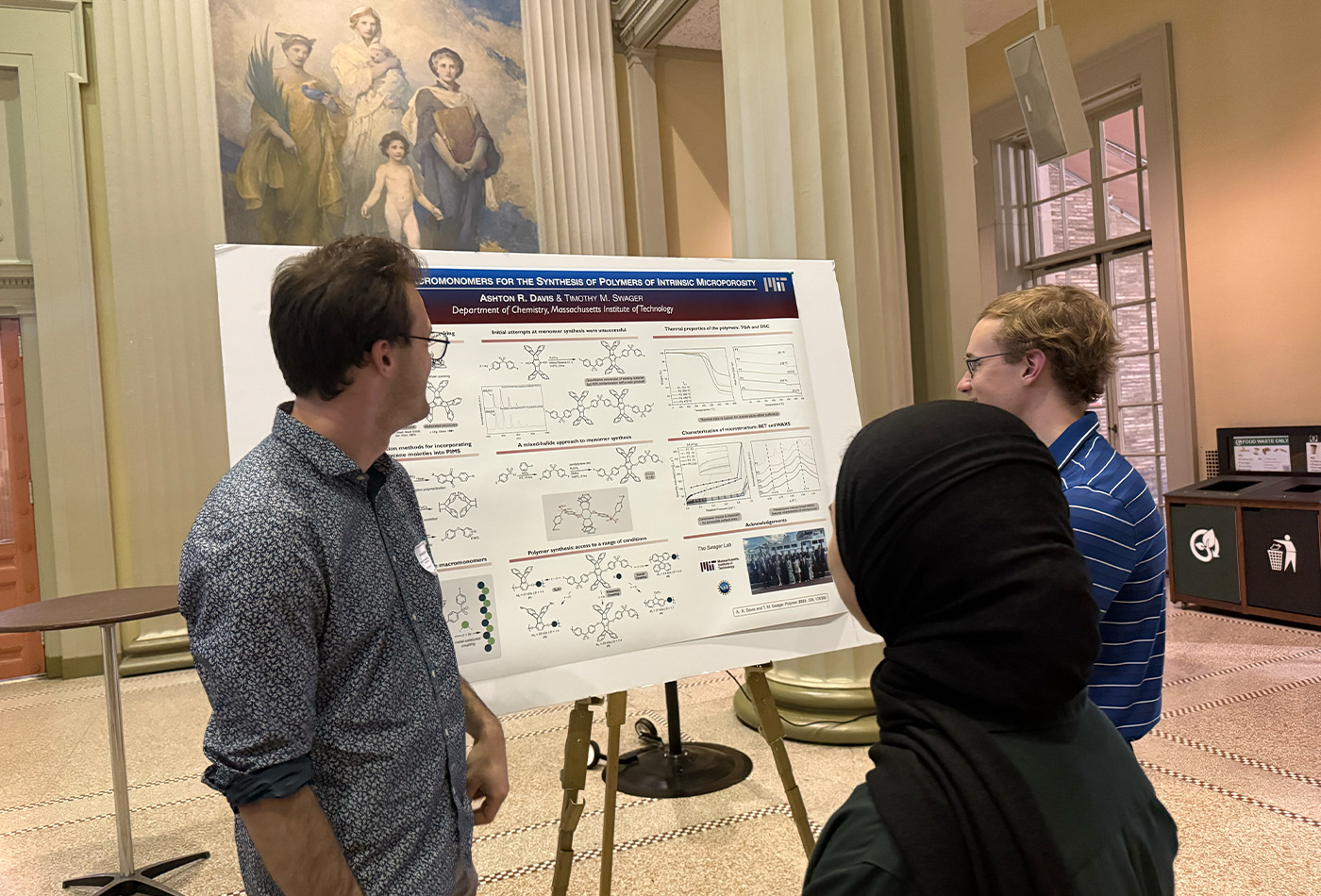 Ashton Davis stands in front of a poster and indicates toward it as Thomas and Mayar look on.