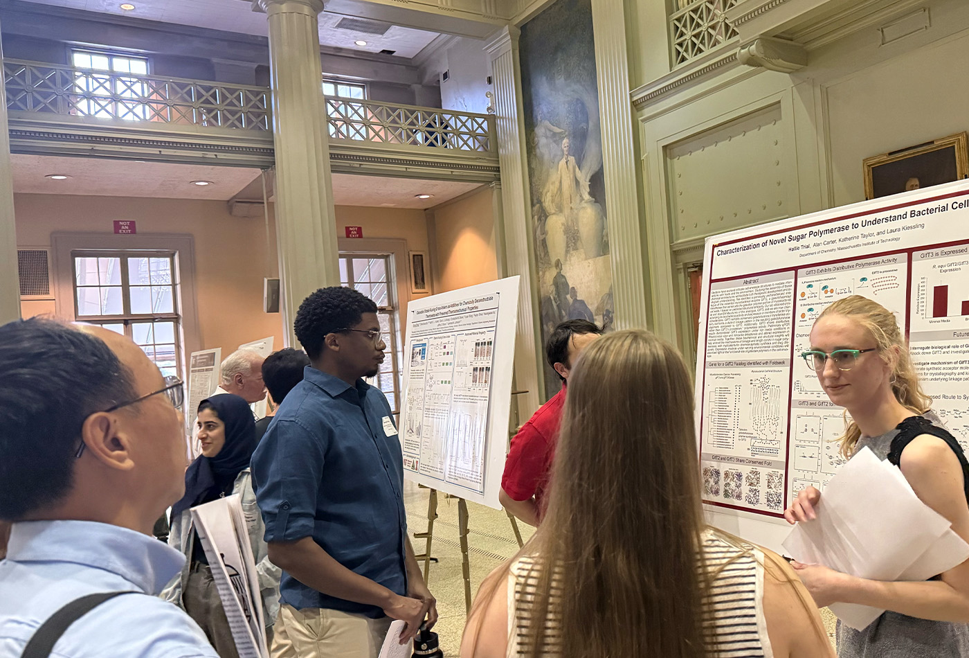 Hallie Trial stands in front of a poster and indicates toward it as Gerard Porter looks on.