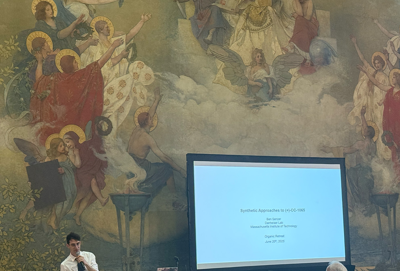 Benjamin Senzer stands in front of a wall and a projection screen.