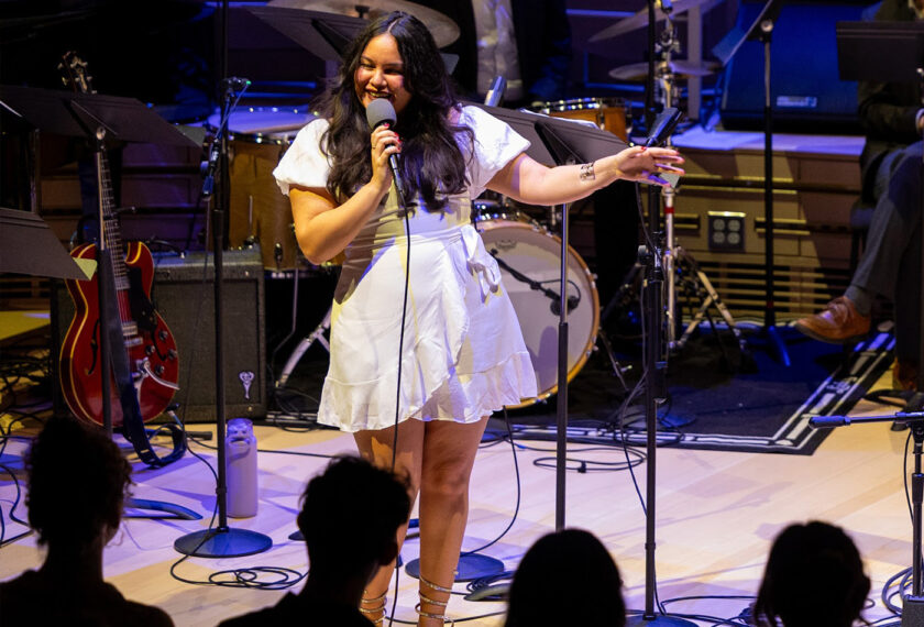 Andrea Delgado performs with a band on stage singing into a microphone wearing a white dress.