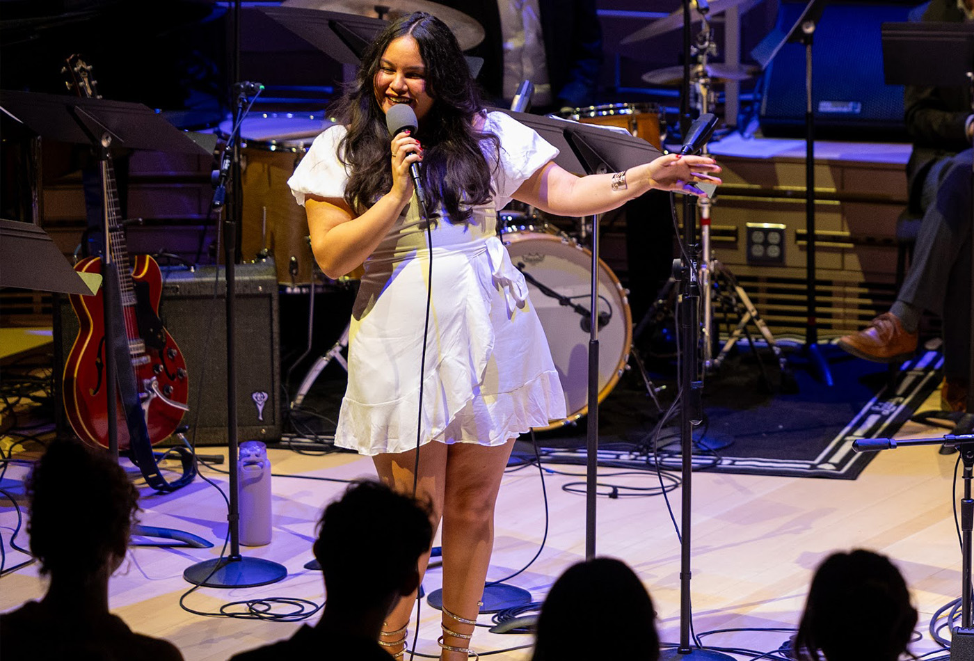 Andrea Delgado performs with a band on stage singing into a microphone wearing a white dress.