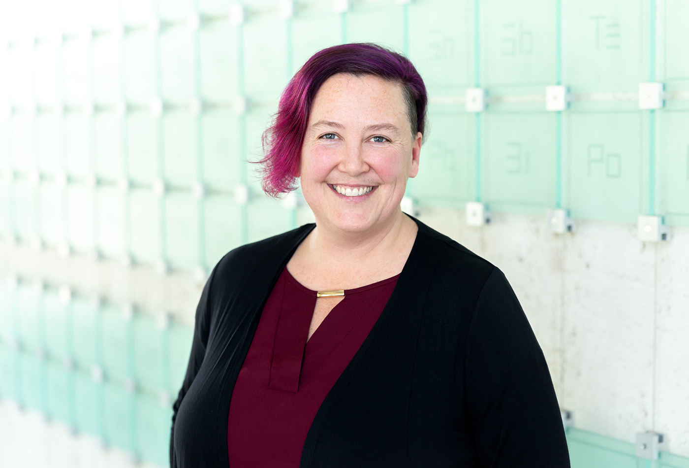Meg Fuchs smiles in front of a glass sculpture of the periodic table of elements.