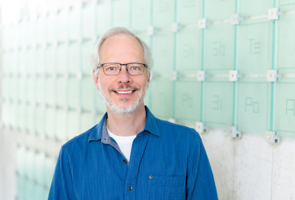 Rick Kayser smiles in front of a glass sculpture of the periodic table of elements.