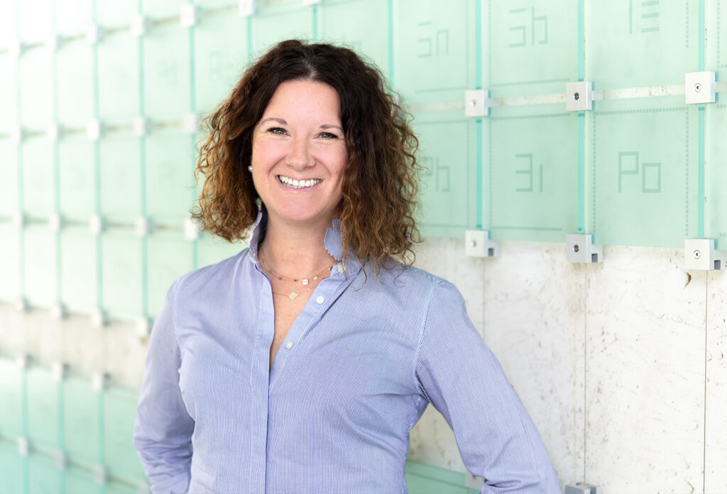 Corrie Lefebvre smiles in front of a glass sculpture of the periodic table of elements.