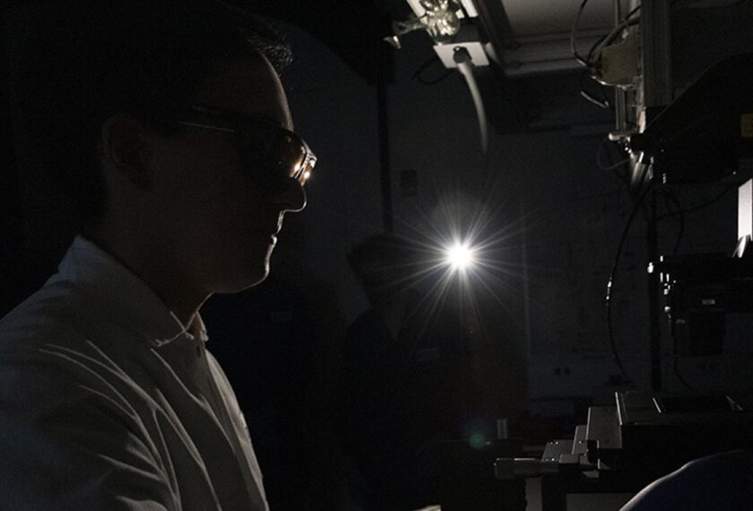 A lab member glances through a mircoscope in a darkened room.