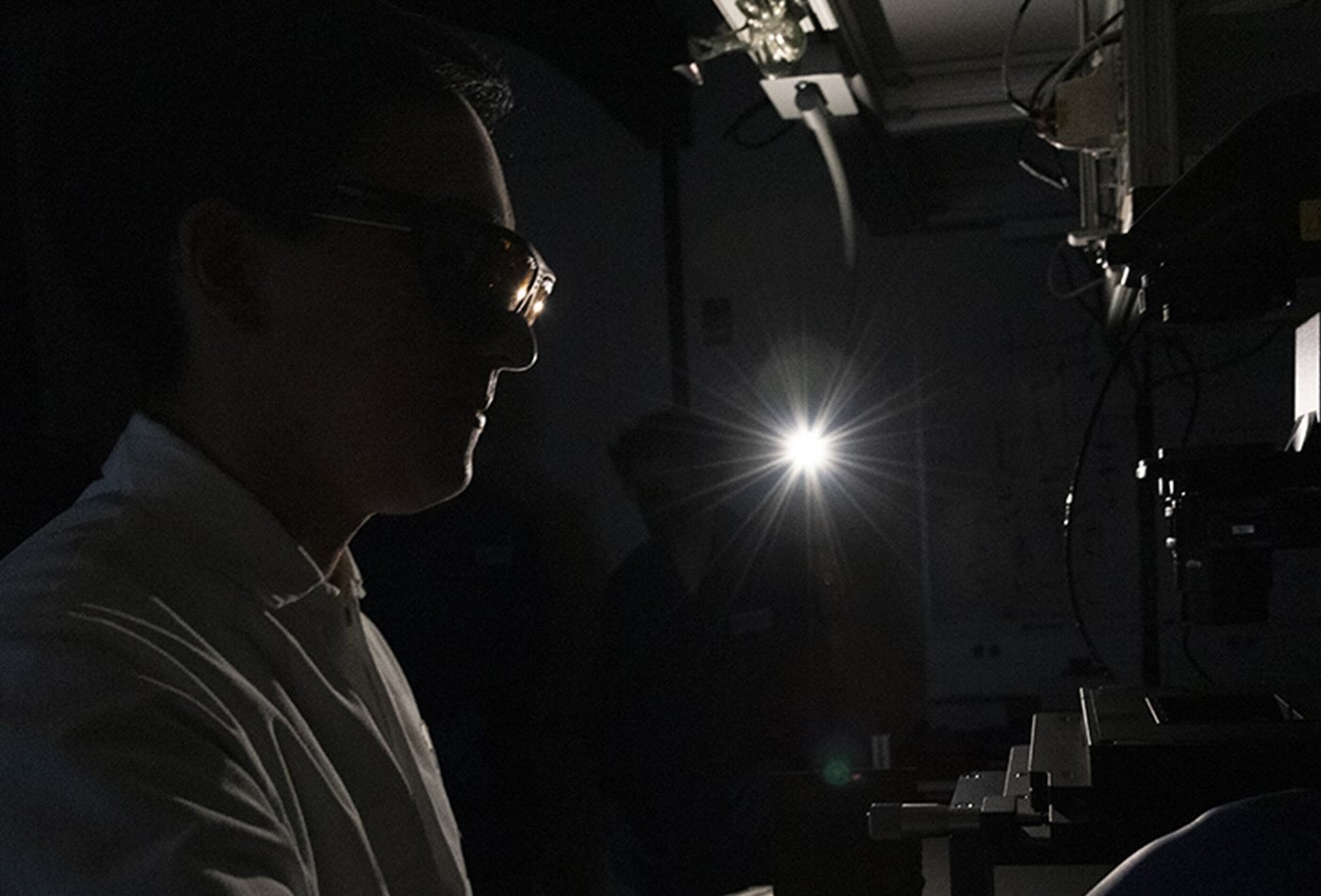 A lab member glances through a mircoscope in a darkened room.